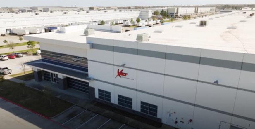 Aerial view of a large industrial building with a logo on the side, located in Houston, Texas. This custom cable manufacturing facility is surrounded by a parking area, with other similar buildings visible in the background.